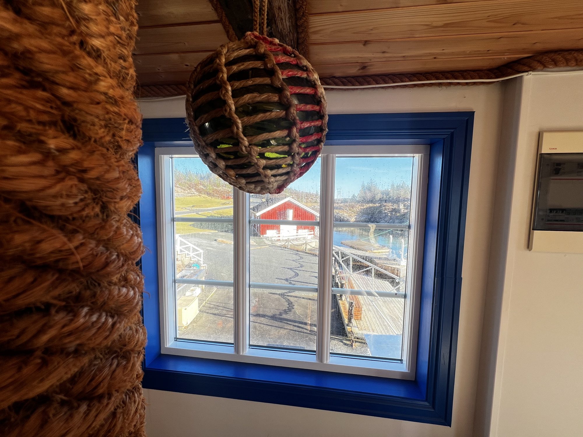 The Heritage — glass fishing float at the blue-framed window with the red boathouse beyond